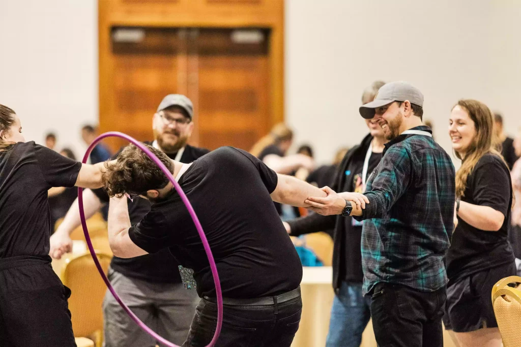 A corporate team stands in a circle and hold each other's arms while passing along a hula hoop without letting go