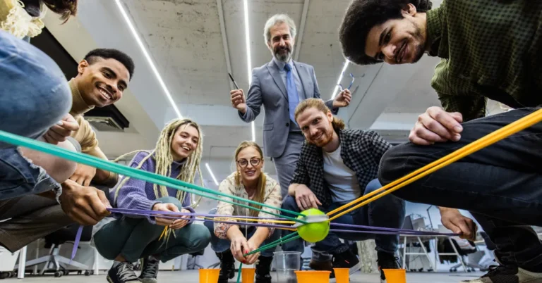 A diverse group of coworkers smiling and playing a teamwork game with strings and a tennis ball, guided by a facilitator in an office setting.