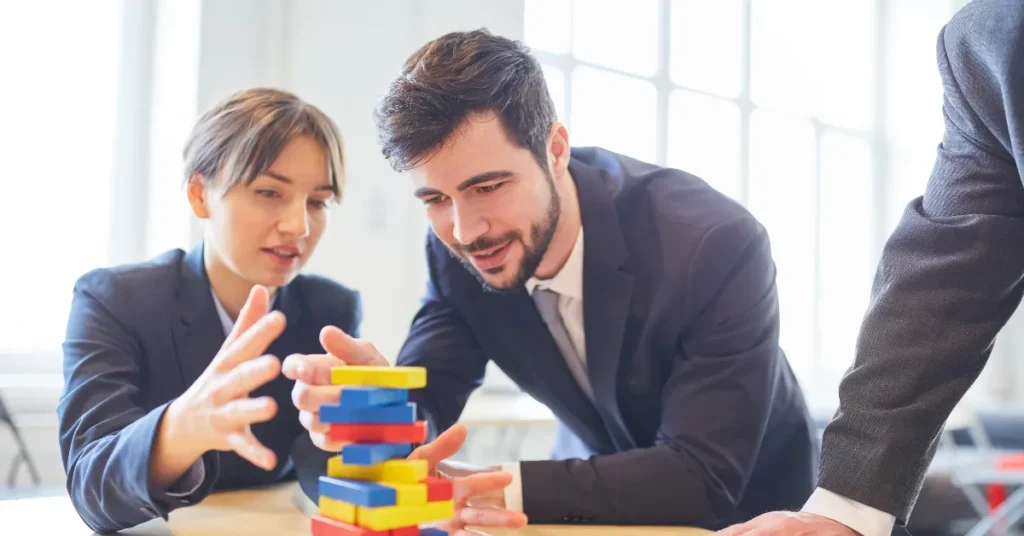 Two business professionals in suits collaborating on a colorful block-stacking teamwork challenge at a table.