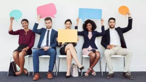 Team of employees sitting in a row holding colorful speech bubbles representing creative employee recognition day ideas to celebrate workplace success.